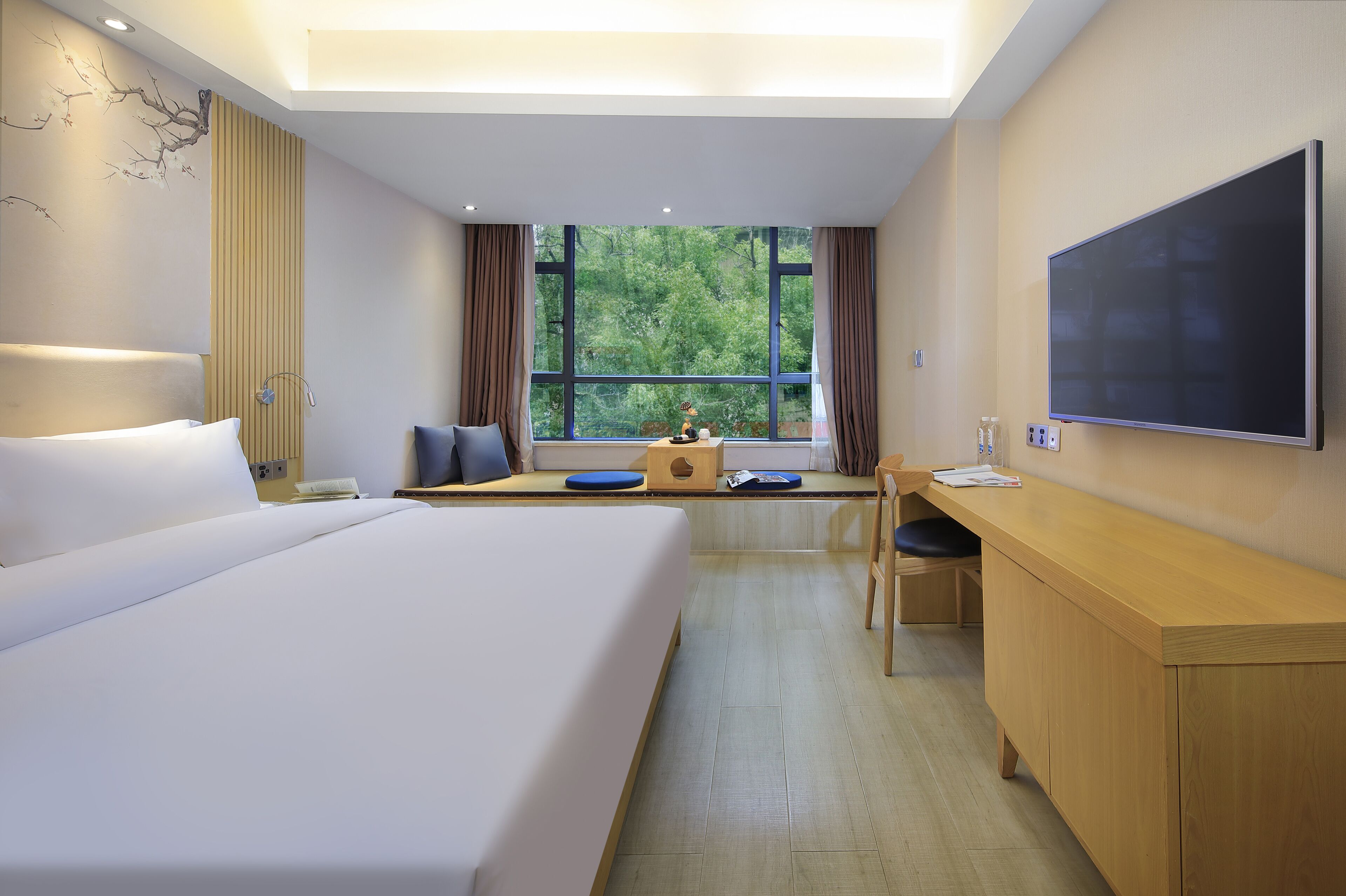 A hotel room with a large bed, a desk, a chair, a flat-screen TV, and a window with a view of trees.