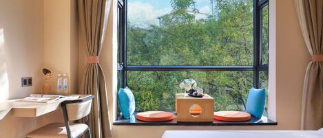 A hotel room with a large window, a desk with a chair, a bed, and a small table with decorative items.