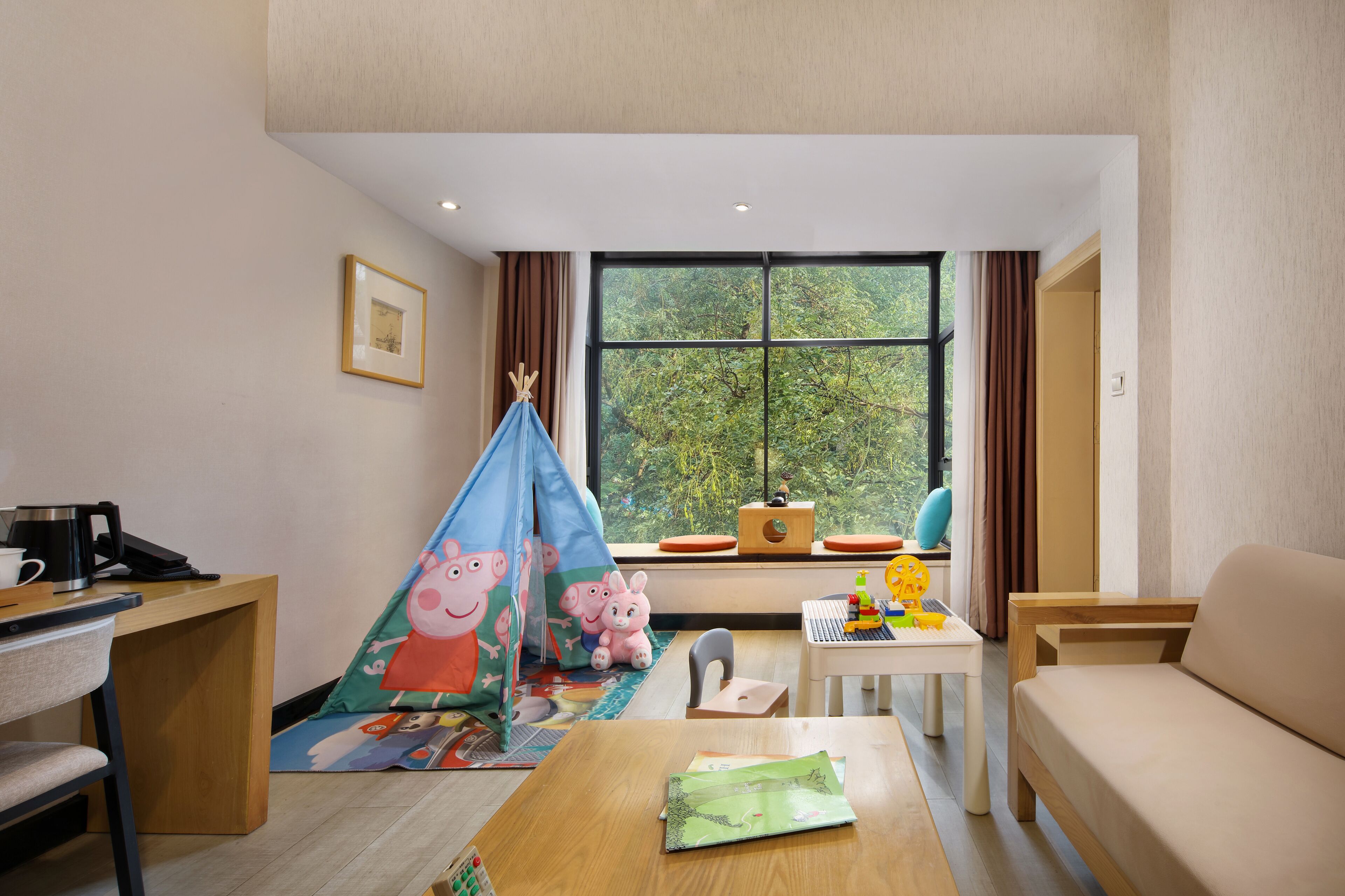 A hotel room with a desk, a sofa, a window with a view of trees, a small table with toys, and a tent with cartoon characters.