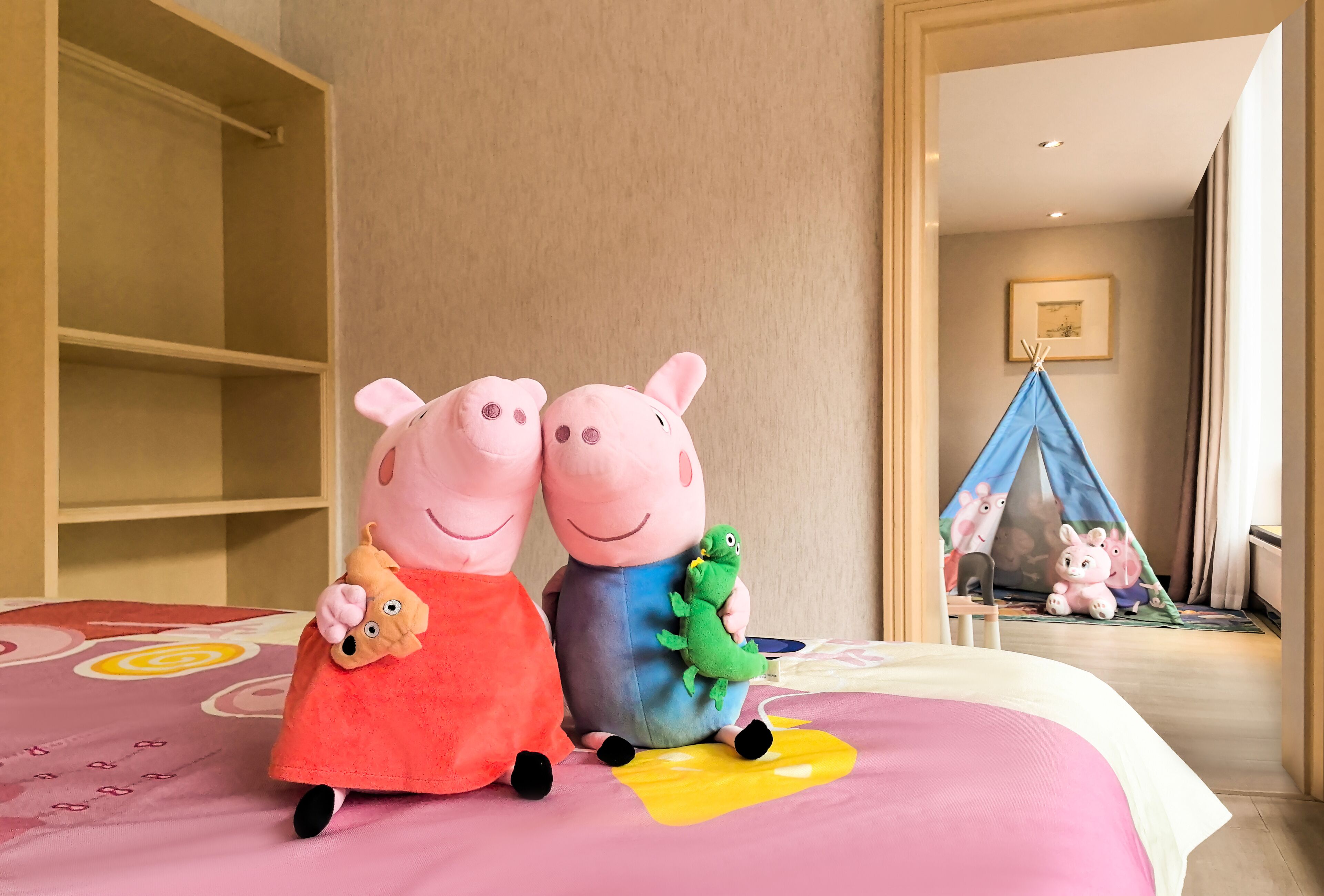 Dos peluches sobre una cama con una colcha rosa y amarilla, un armario de madera y una tienda de campaña azul al fondo.