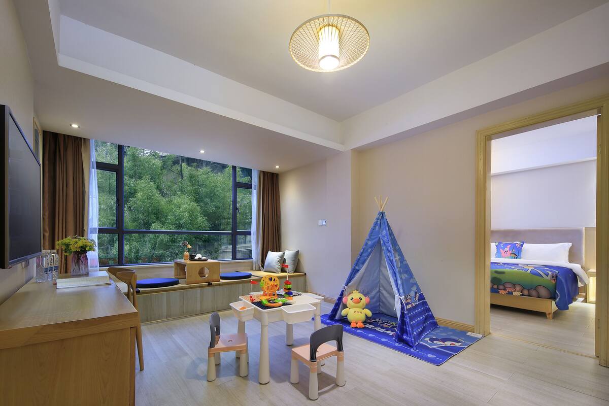 A child-friendly room with a large window, a desk with toys, a bed with a blue bedspread, and a tent.
