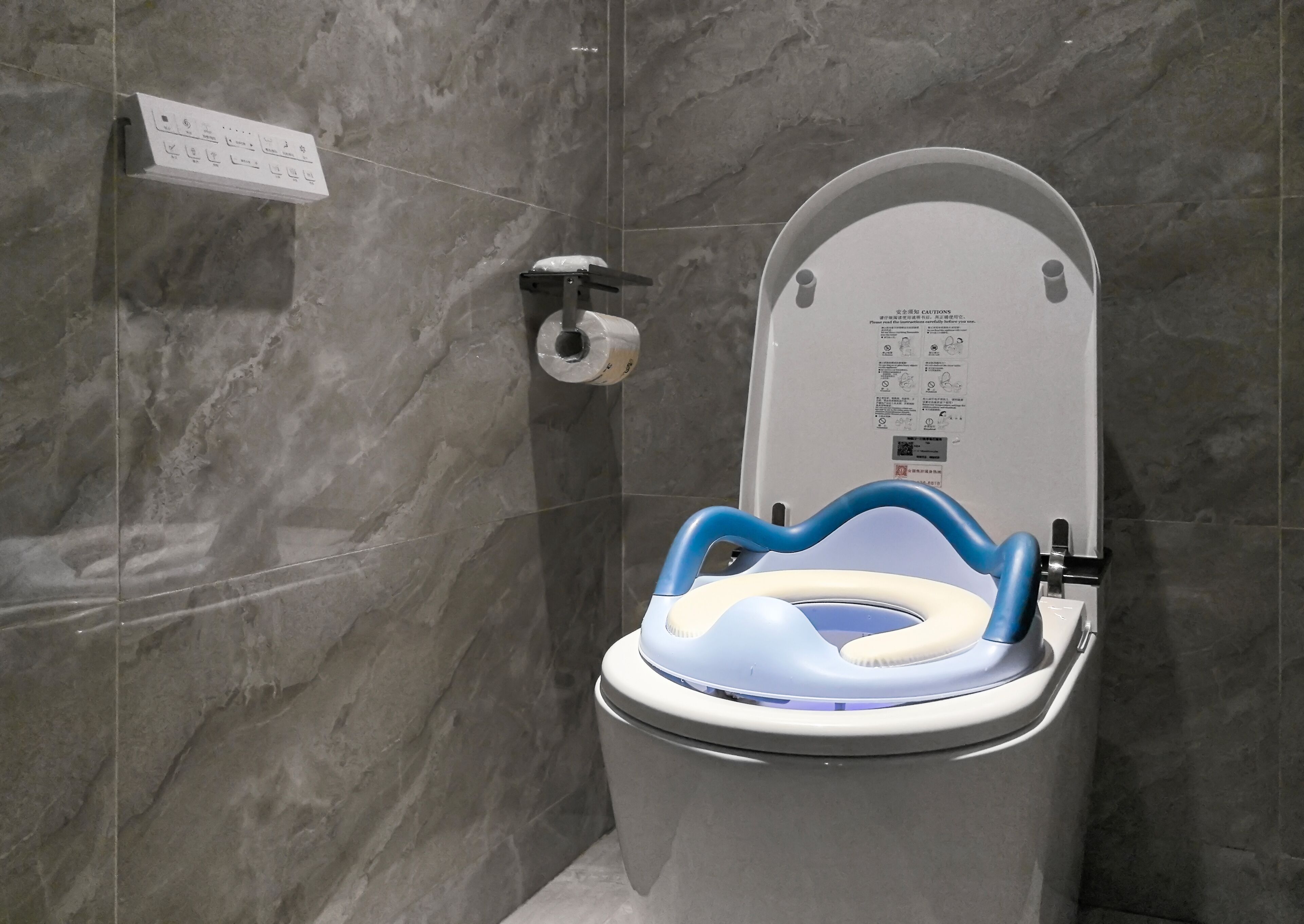 Un inodoro moderno con una protección azul de seguridad, un dispensador de papel higiénico y un letrero de precaución en la pared.