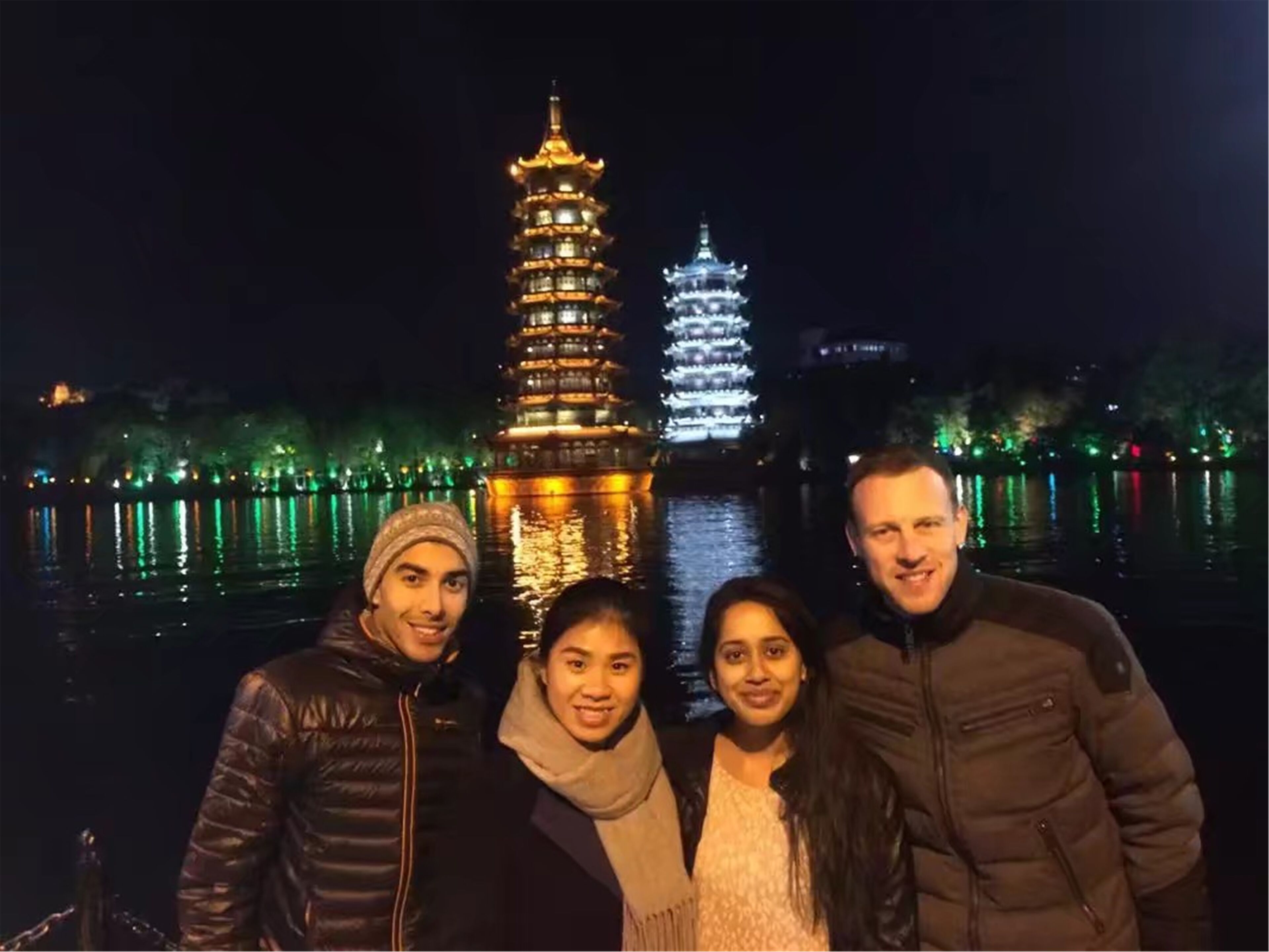 Um grupo de pessoas em frente a um pagode tradicional com vários níveis, iluminado por luzes, e um corpo d'água refletindo as luzes.