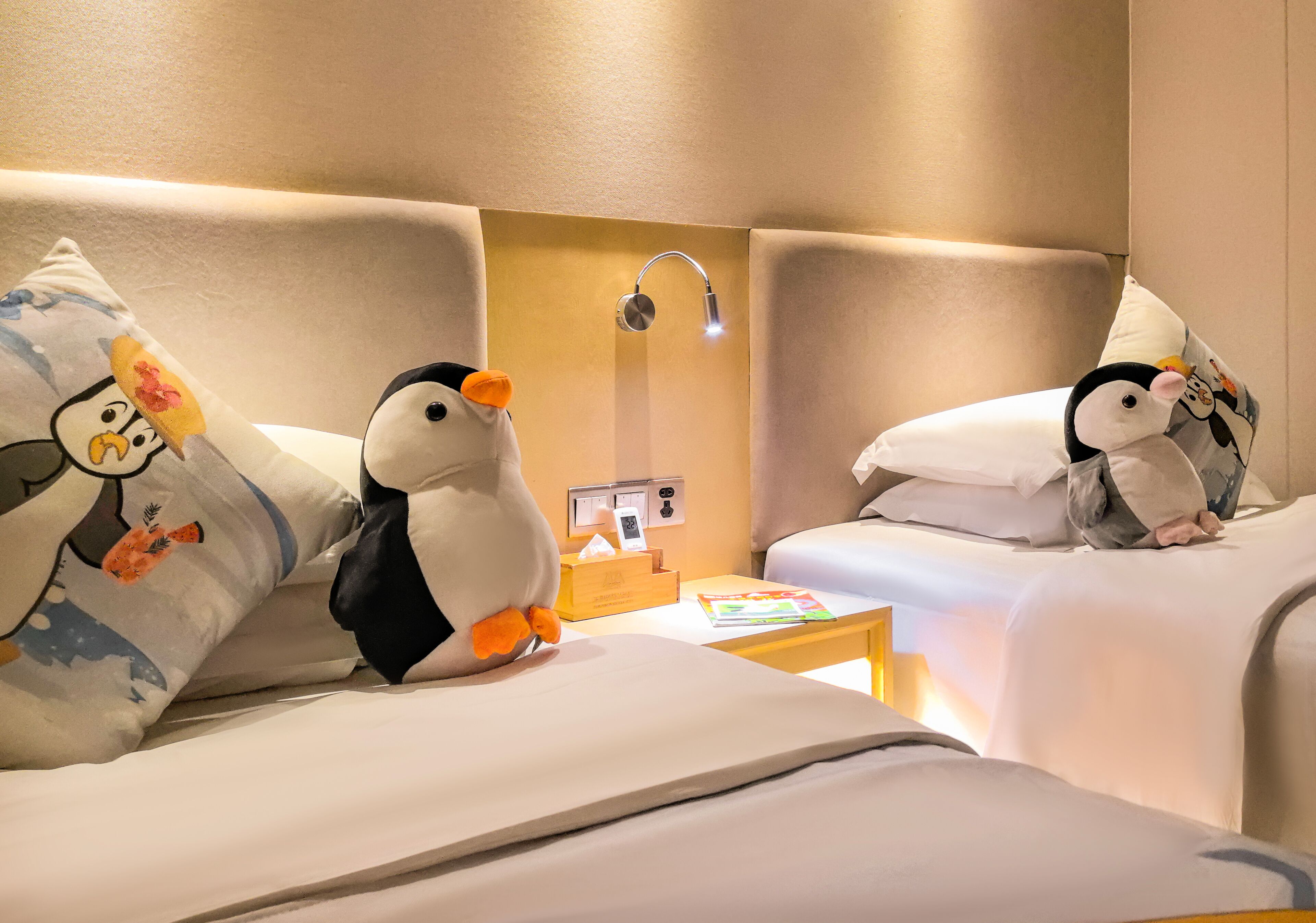 A hotel room with two beds, each with a plush penguin toy and a pillow with a cartoon character. There is a nightstand with a lamp, a phone, and a small table with a book and a tissue box.