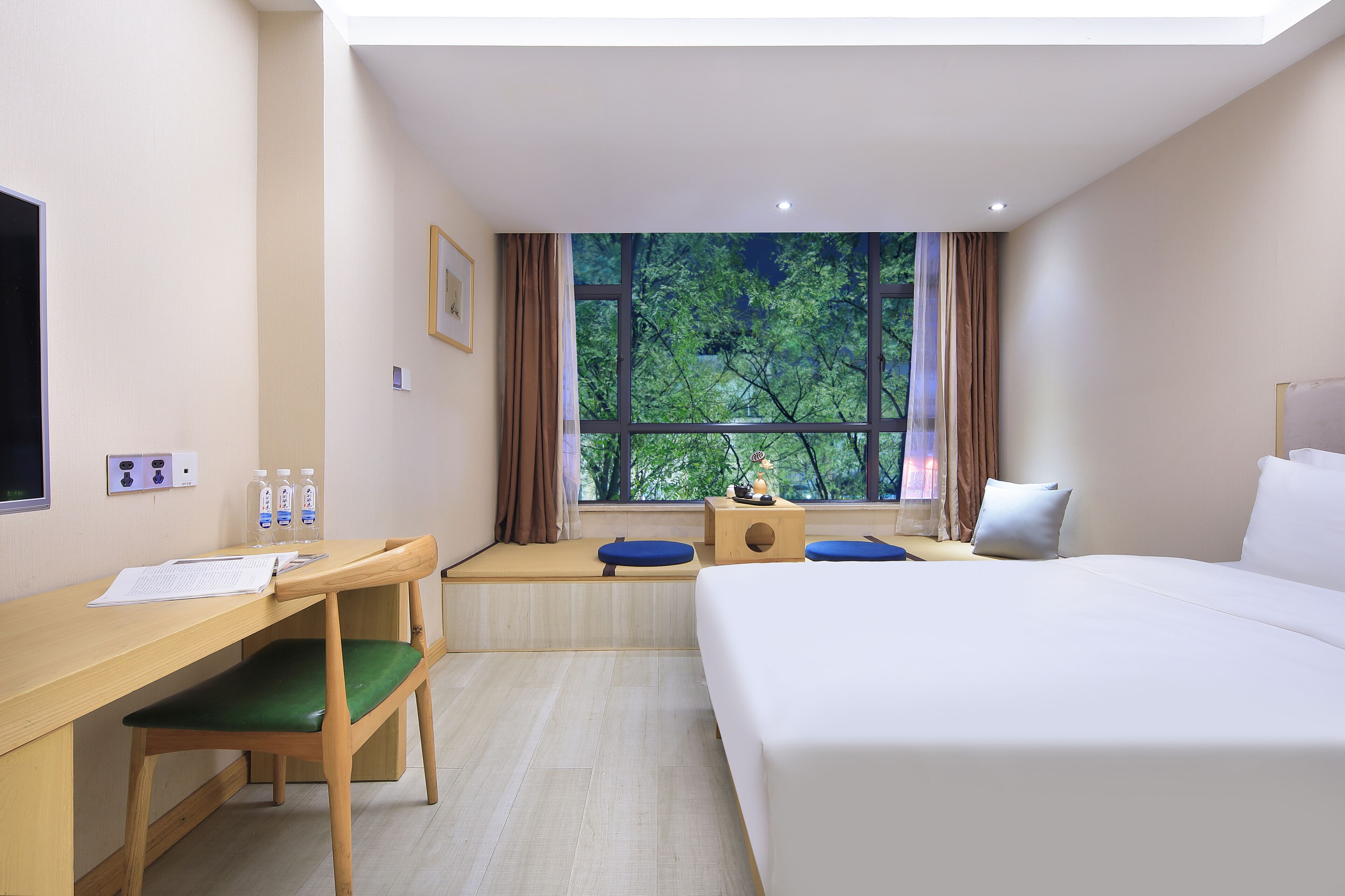 A hotel room with a bed, a desk, a chair, a window with a view of trees, and a small table with a lamp.