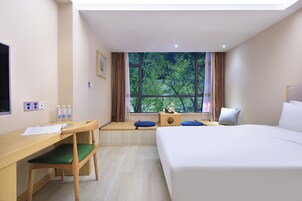 Habitación de hotel con cama, escritorio, silla, ventana con vista a árboles y una mesita de noche con lámpara.