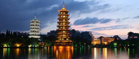 A traditional pagoda with multiple tiers, illuminated at dusk, reflecting on a calm body of water, with modern buildings in the background.
