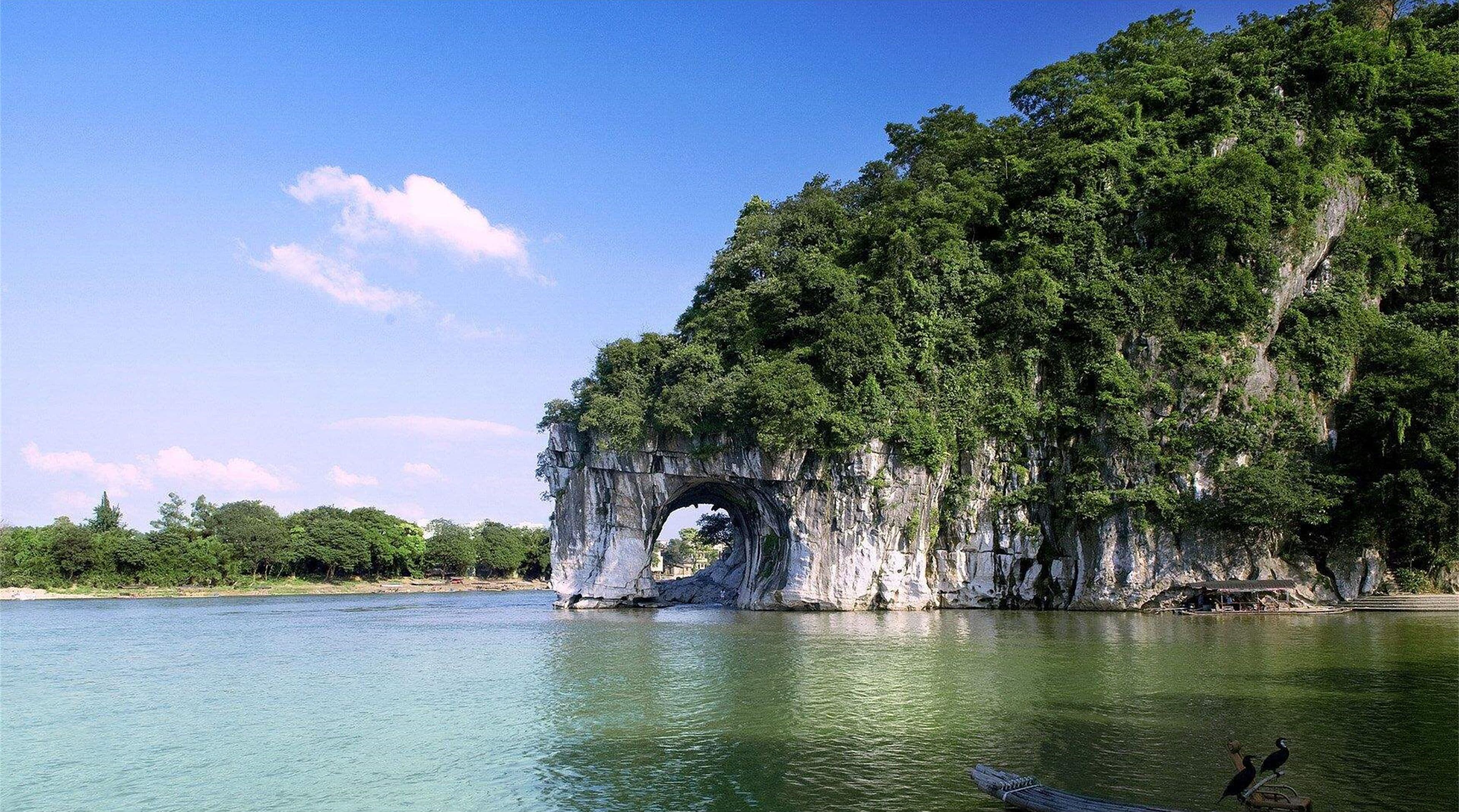 A natural rock formation with a cave-like opening surrounded by lush greenery and a calm body of water.