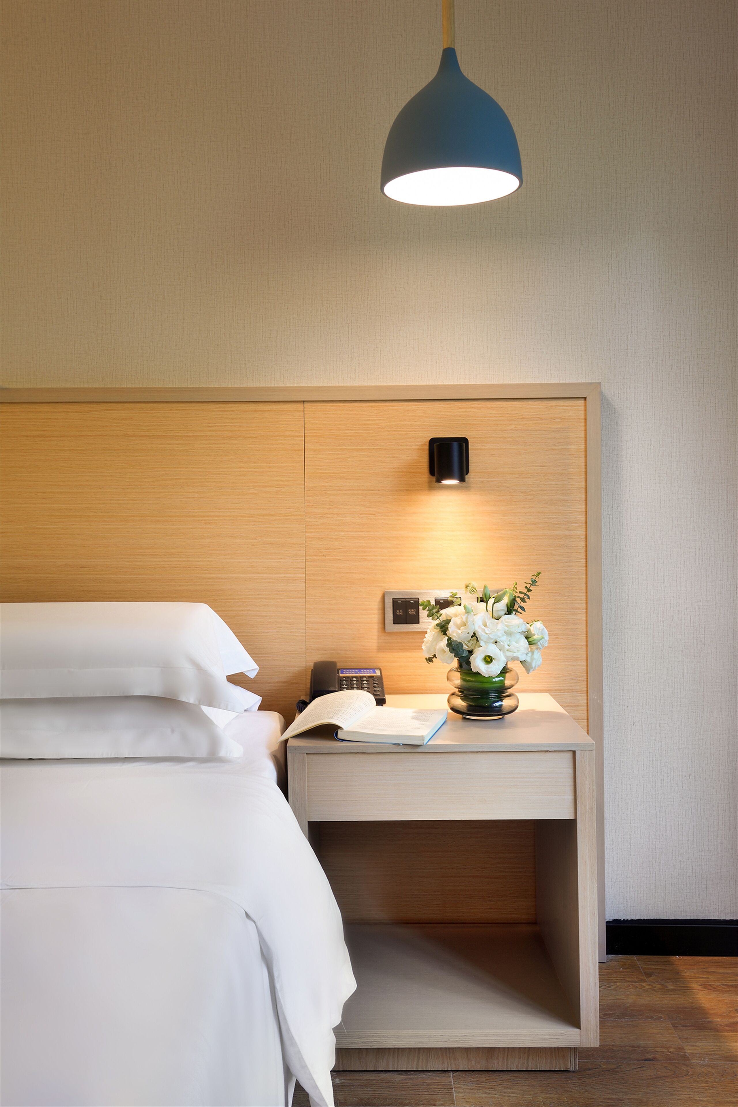 Una cama bien tendida con sábanas blancas, una mesita de noche con un jarrón de flores, una lámpara y un aplique de pared.