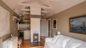 Een gezellige, goed verlichte kamer met een houten vloer, een witte bank, een kitchenette met een fornuis en een schilderij van een zeilboot aan de muur.