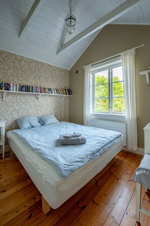 A bedroom with a wooden floor, a bed with a striped bedspread, a bookshelf with books, a window with curtains, and a white radiator. - Vaxholm Harbour Bed and Experiences (Vaxholm)