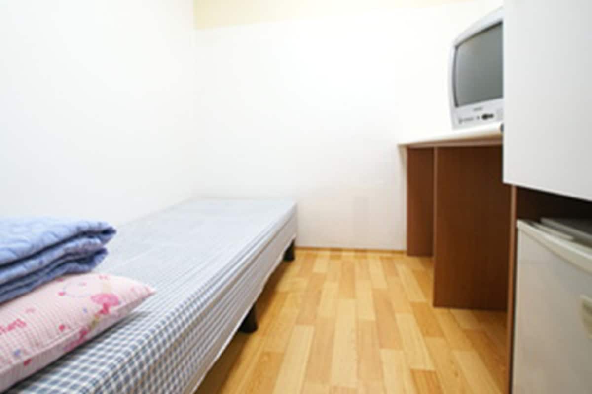 A small, single-bed room with a wooden floor, a bed with a blue and white checkered bedspread, and a television on a wooden cabinet.