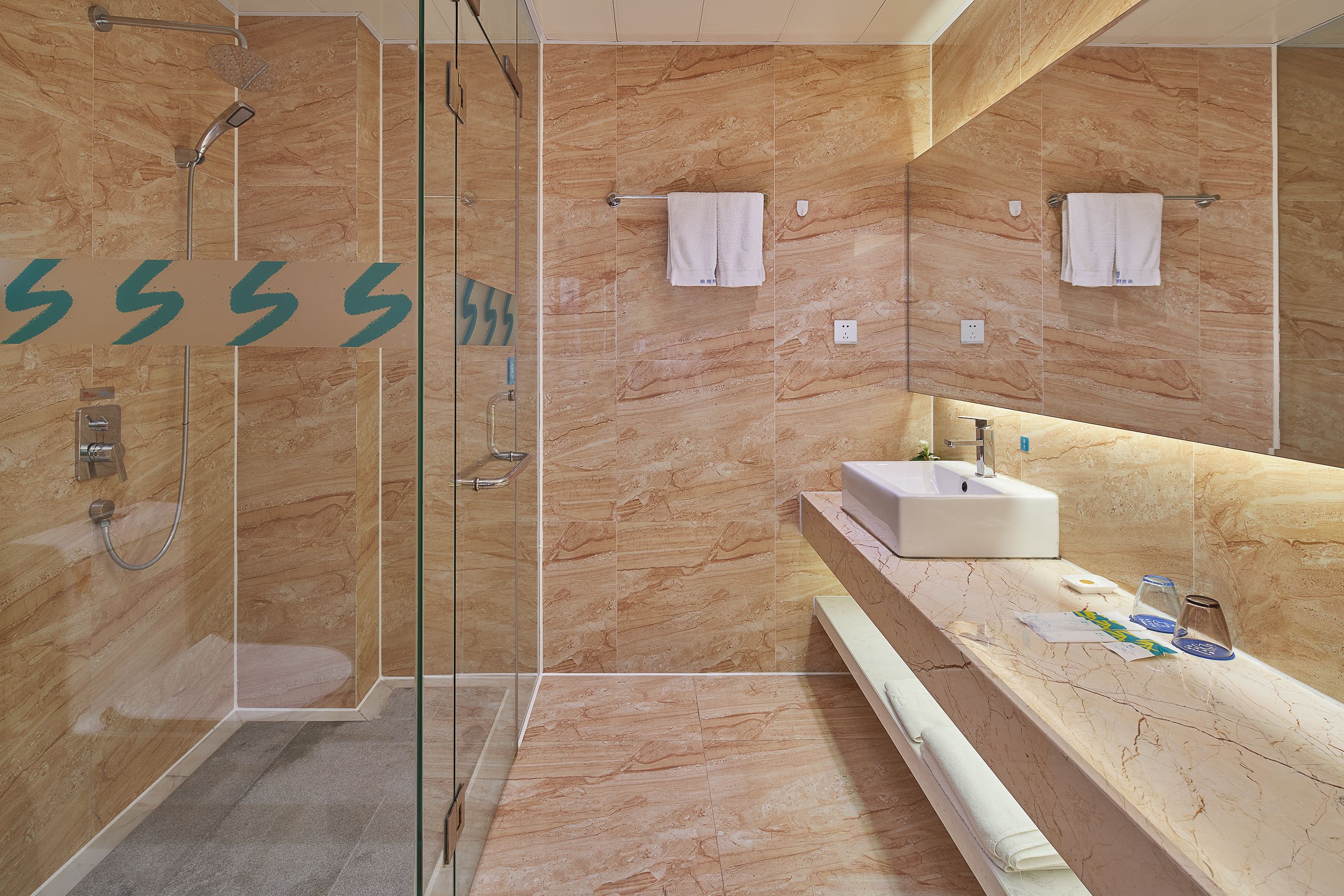 A modern bathroom with a glass-enclosed shower, marble walls, a sink, and two towels hanging on hooks.
