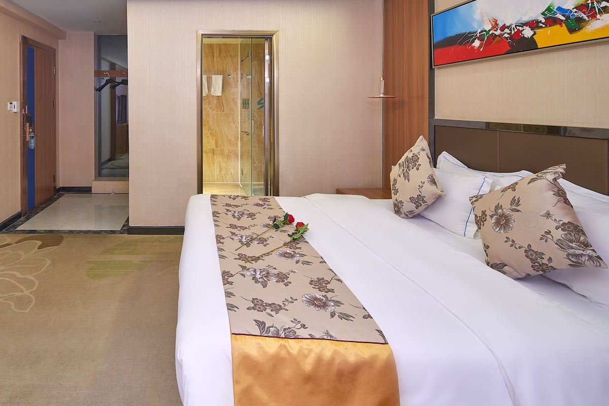 A hotel room with a bed, a television, a shower, and a hallway leading to another room.