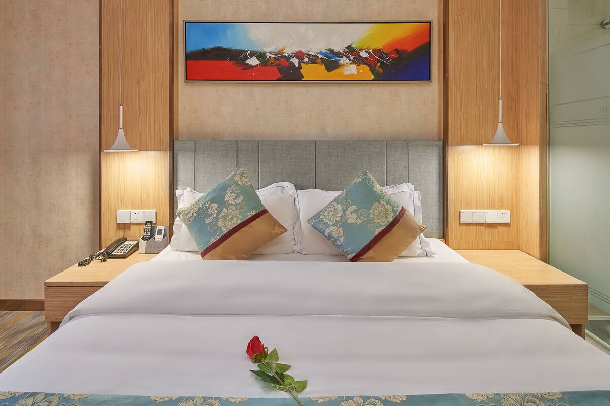 A hotel room with a large bed, two bedside tables, a mounted painting, and a single red rose on the bed.