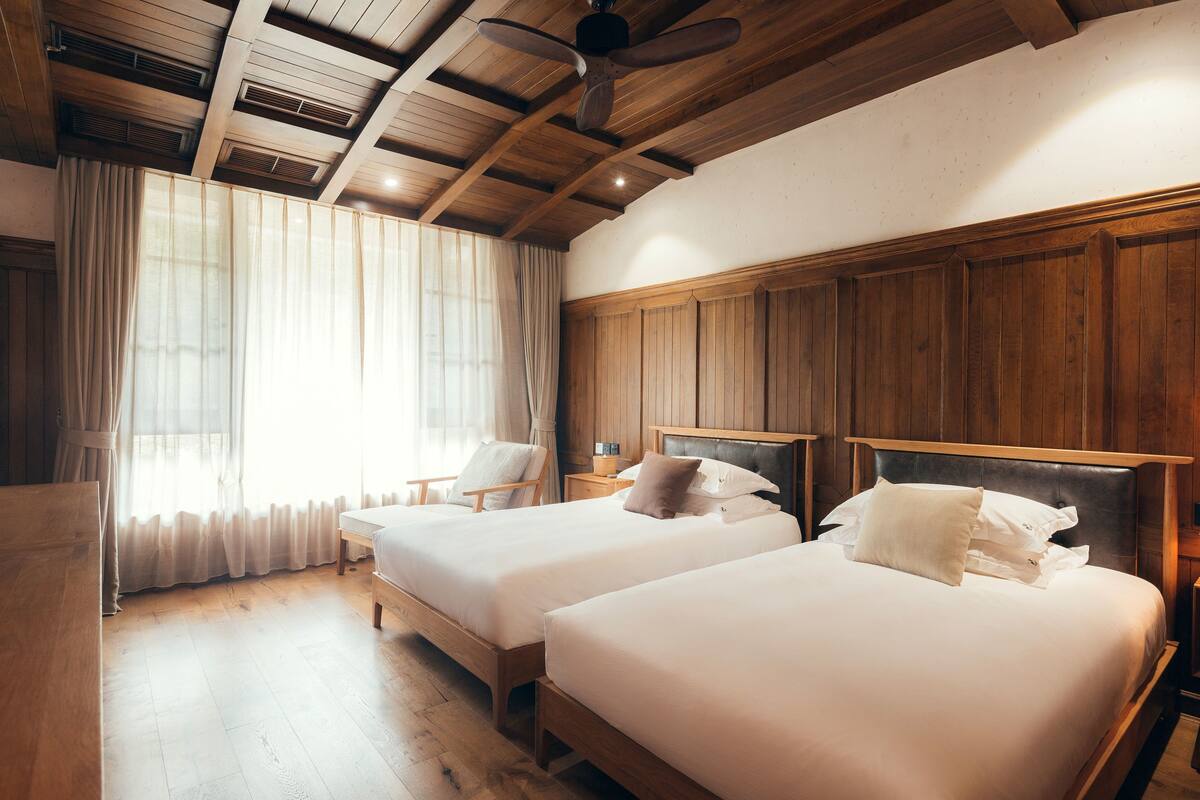 Ein Zimmer mit zwei Betten, Holzbalkendecke und einem großen Fenster mit Vorhängen.