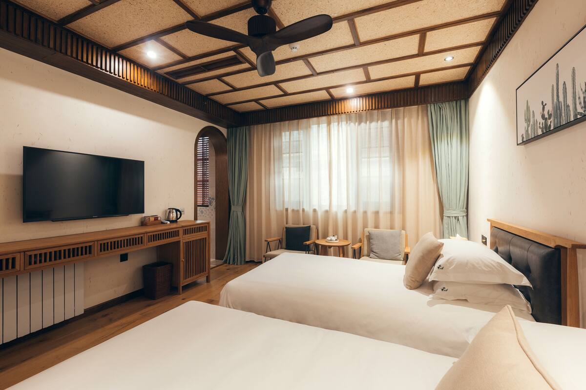 Ein Hotelzimmer mit zwei Betten, einem Fernseher, einem Deckenventilator, einem Fenster mit Vorhängen und einem Gemälde an der Wand.