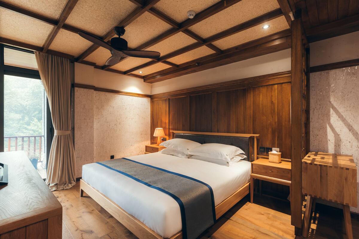 Ein Hotelzimmer mit Holzboden, einem Bett mit blauen Akzenten, einem Nachttisch aus Holz und einem Fenster mit Vorhängen.