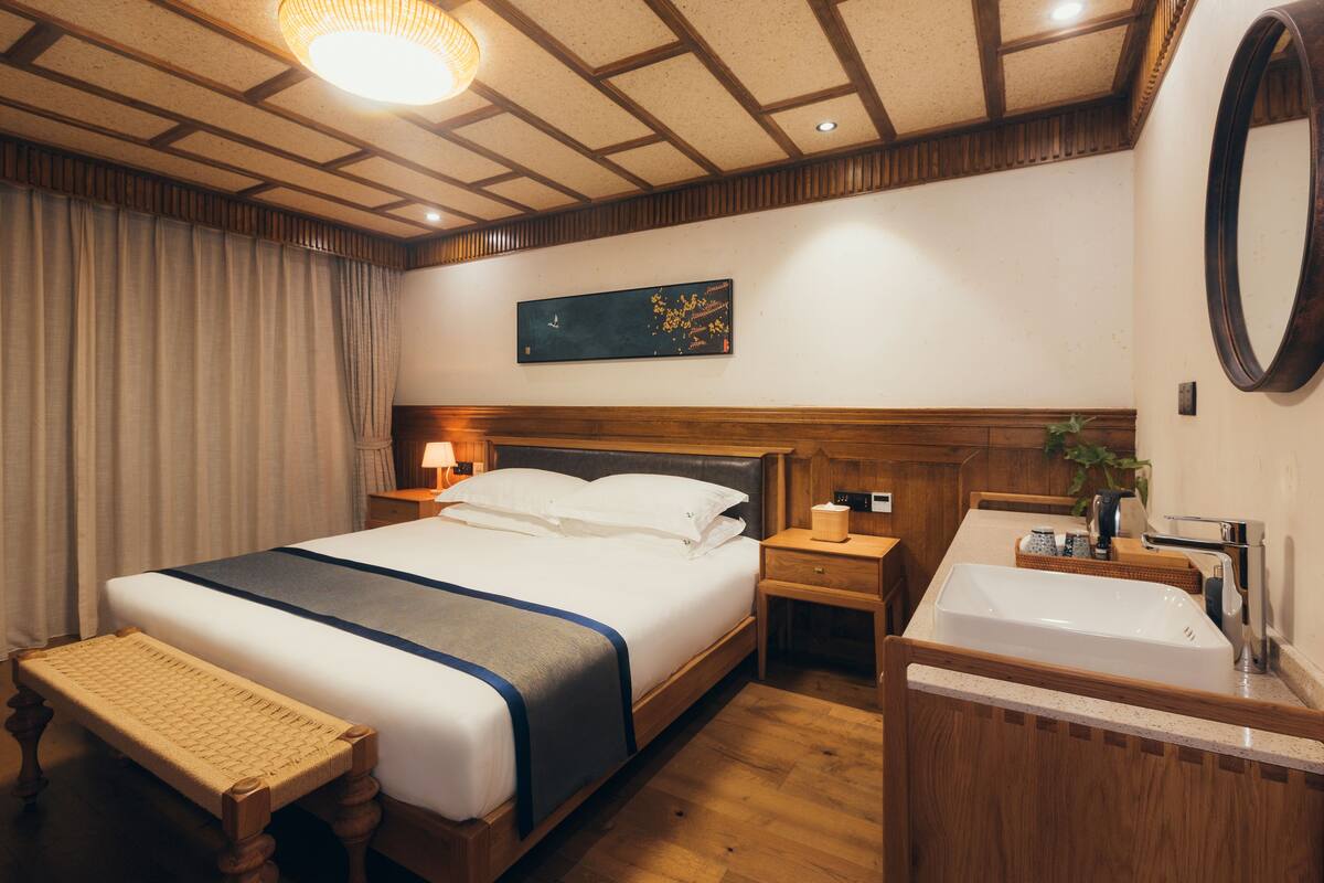 Ein Hotelzimmer mit einem großen Bett, einem Holzkopfende, Nachttischen, einer Bank, einem Badezimmer mit Waschbecken und Spiegel sowie einem Bild an der Wand.