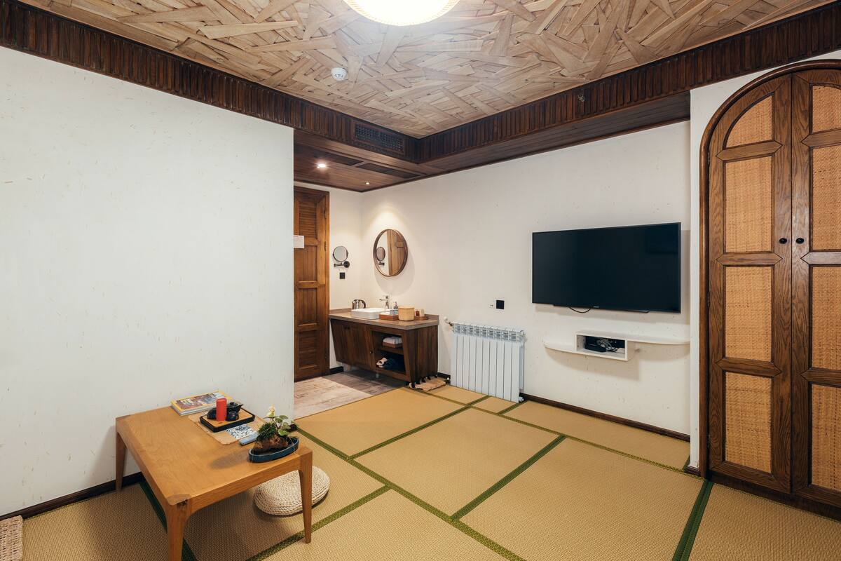 Ein Zimmer mit Tatami-Böden, einem Holztisch, einem an der Wand montierten Fernseher und einer traditionellen japanischen Tür.