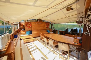 A boat's deck with a wooden table, chairs, life rings, and a steering wheel.