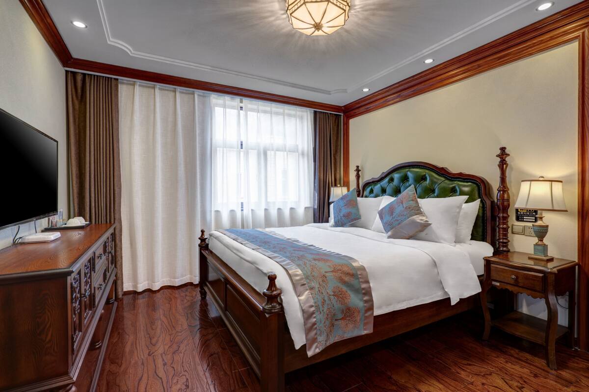 Une chambre d’hôtel avec un grand lit, une tête de lit en bois, un téléviseur posé sur une commode en bois et une table de chevet avec une lampe.