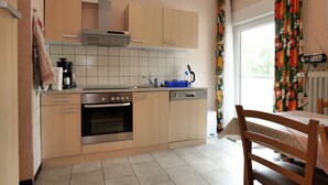 Una cocina compacta con estufa, horno y lavavajillas, una mesa de comedor con una silla y un ventanal con cortinas.