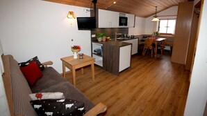 Ein gemütliches Wohnzimmer mit Holzbalkendecke, einem an der Wand montierten Fernseher, einem Essbereich mit Tisch und Stühlen sowie einer Küche mit Schränken und einer Mikrowelle.
