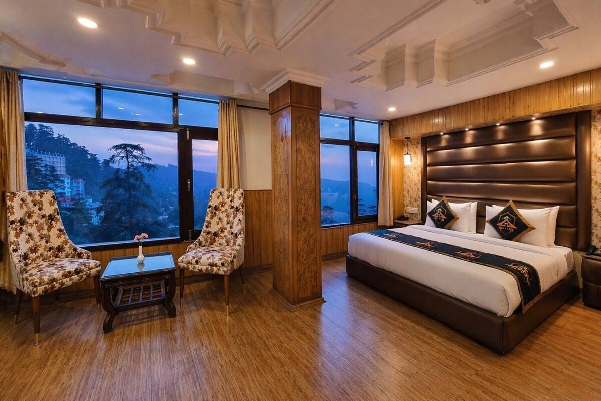 A hotel room with a large bed, two armchairs, a small table, and a view of a mountainous landscape through large windows.