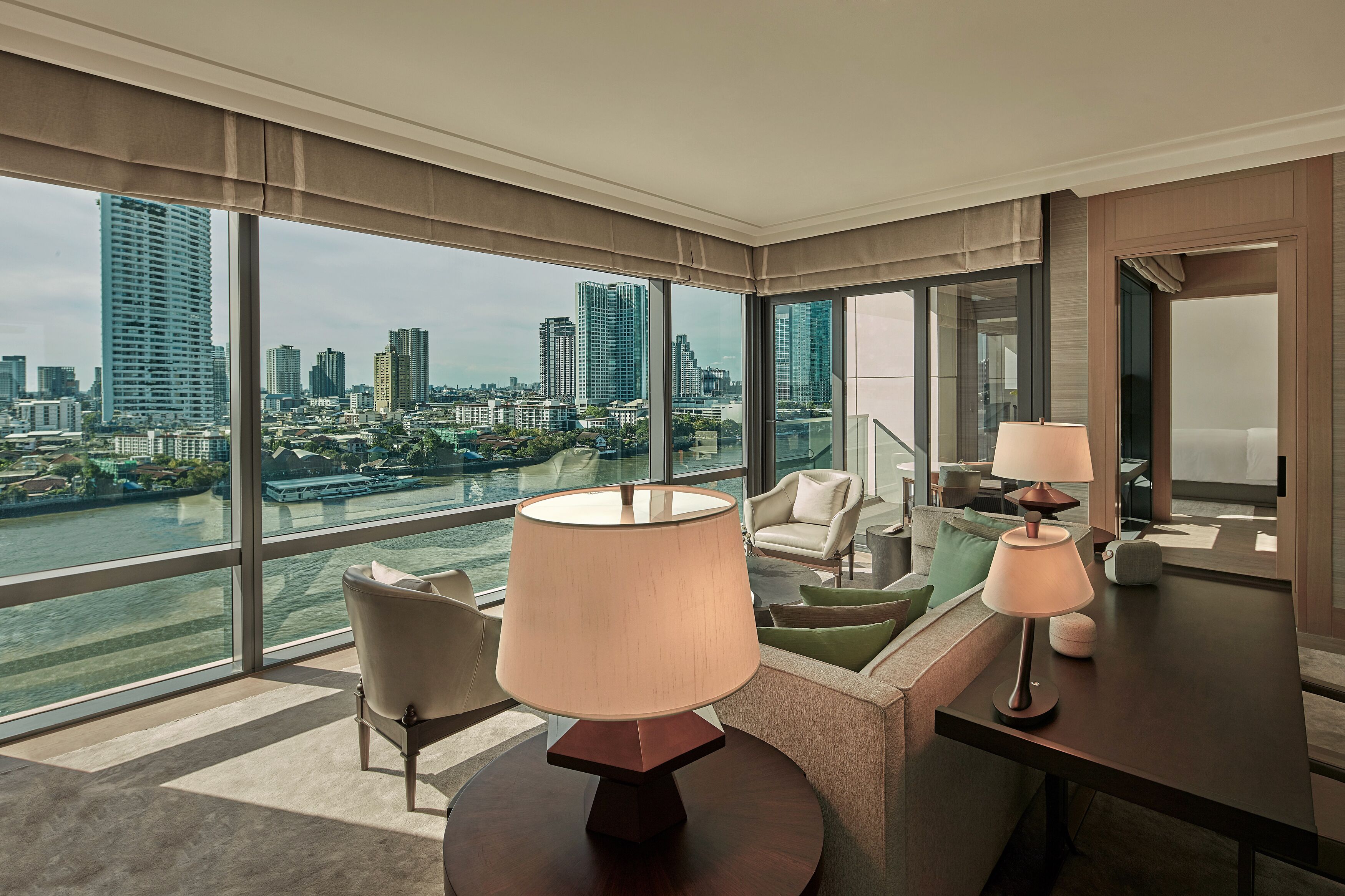 A modern hotel room with a large window offering a city view, a round table with a lamp, and a sofa with cushions.