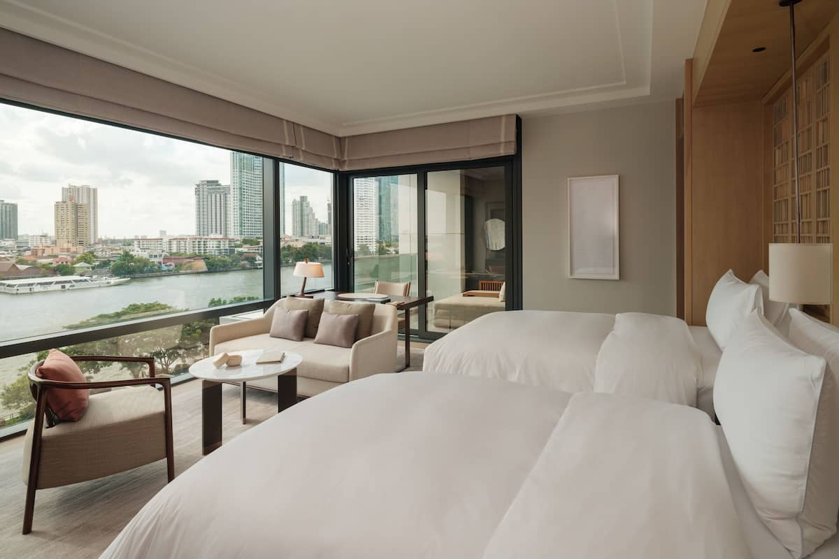 A modern hotel room with a large bed, a seating area with a table, and a view of a cityscape and river.