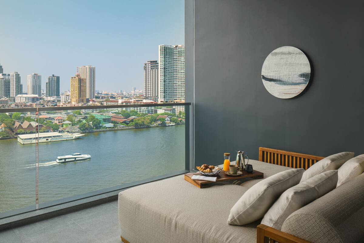 A room with a large window overlooking a river and cityscape, a bed with pillows, a tray with breakfast items, and a round wall decoration.