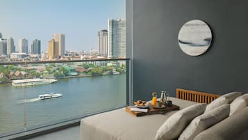 A room with a large window overlooking a river and cityscape, a bed with pillows, a tray with breakfast items, and a round wall decoration.