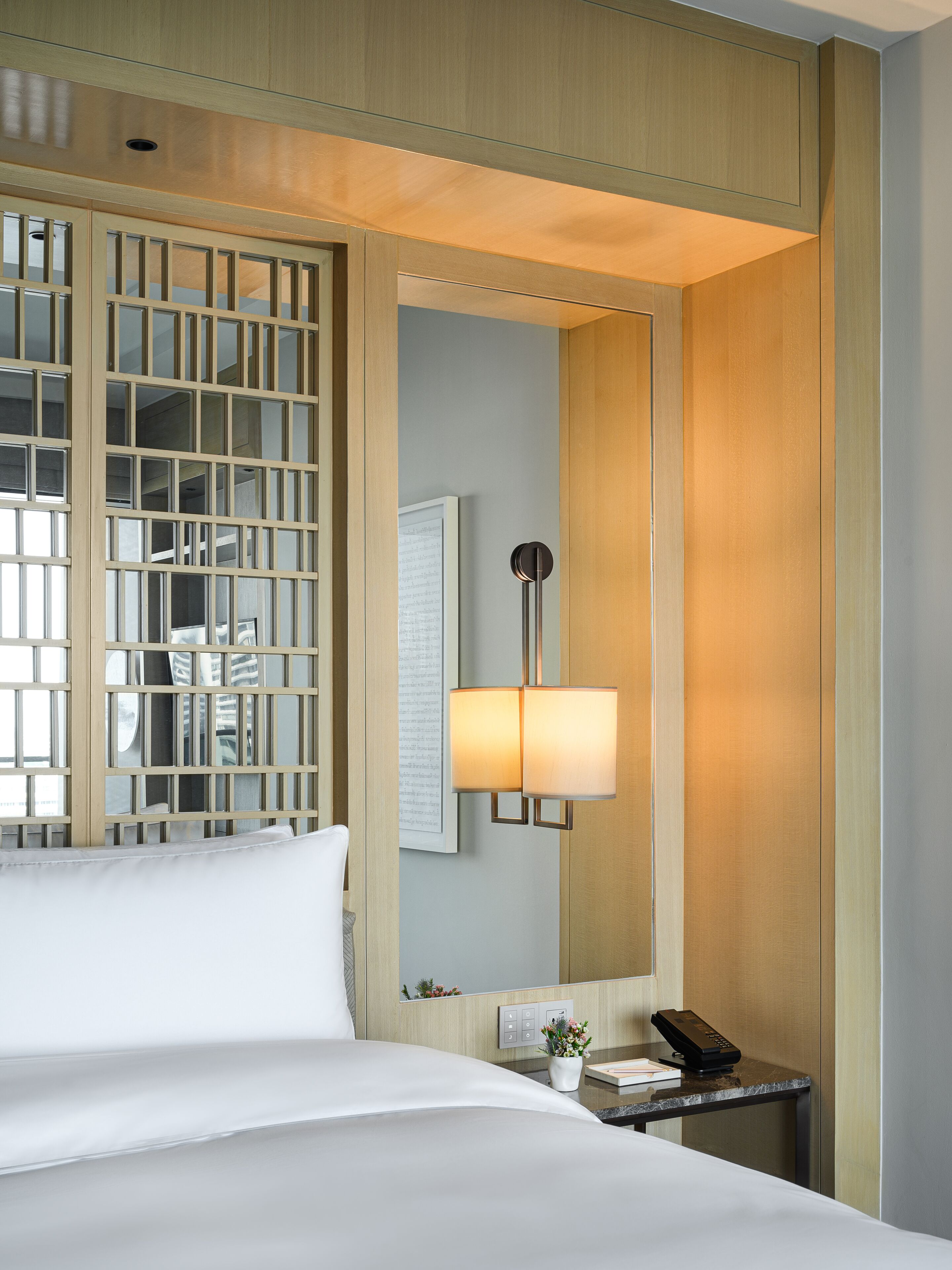 A hotel room with a bed, a nightstand, a lamp, a mirror, and a wooden partition.