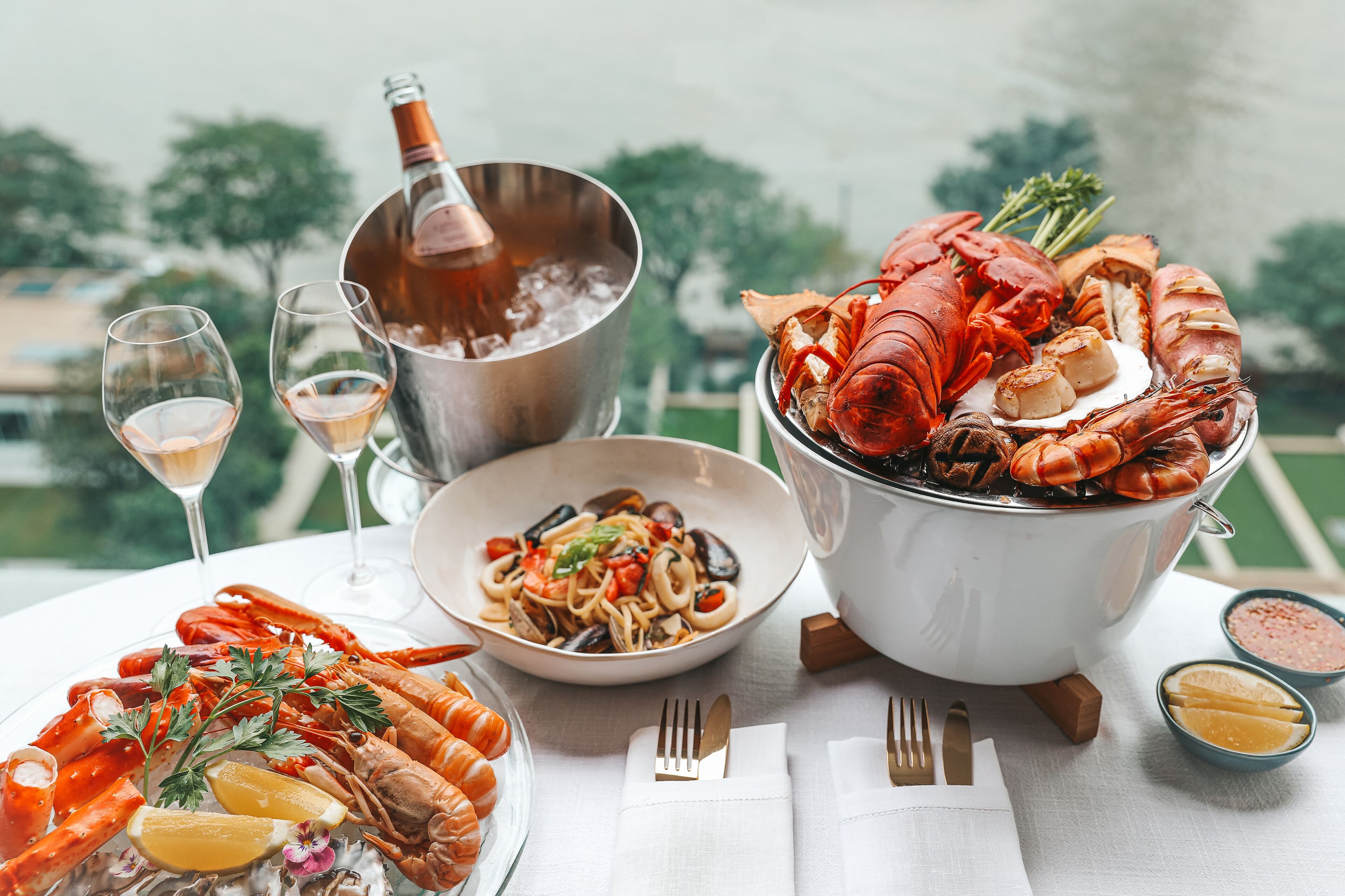 A seafood platter with various shellfish and a pot of lobster, a bowl of pasta, and a bottle of champagne on a table with a view of trees.