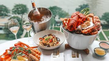 Un plateau de fruits de mer comprenant divers crustacés et un homard entier, un plat de pâtes et une bouteille de champagne, le tout disposé sur une table offrant une vue sur les arbres.
