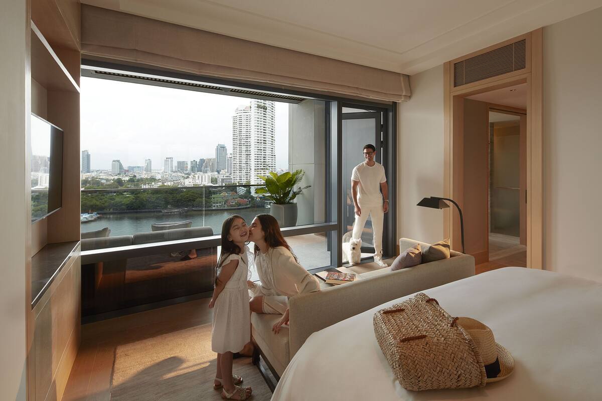 A modern hotel room with a large window offering a city view, a bed with a woven bag, and a sofa with a person standing nearby.