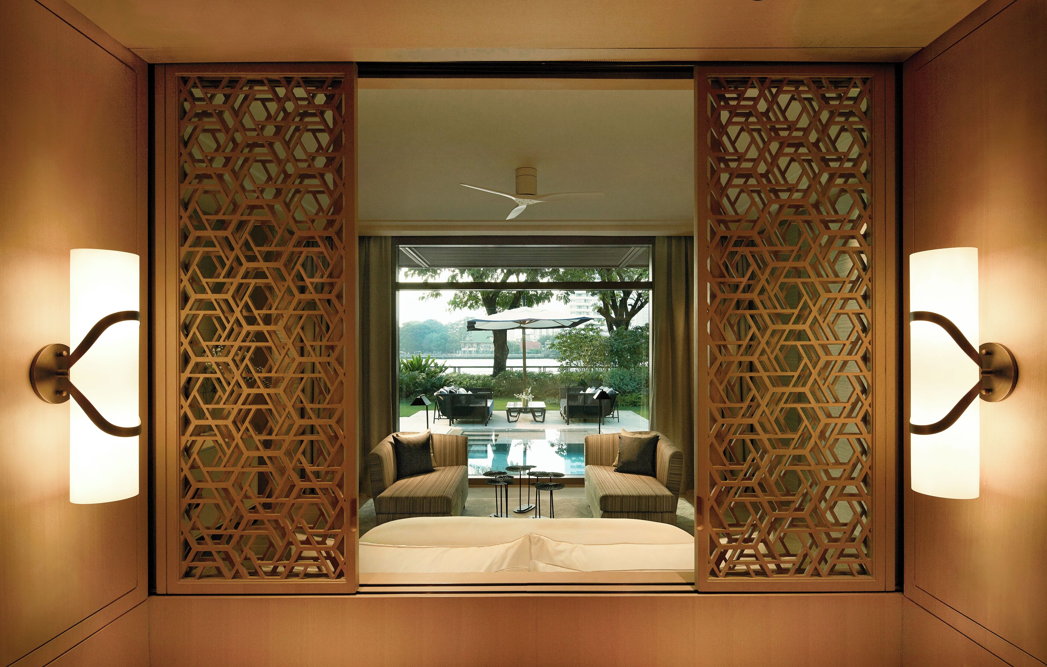 A hotel room with a bed, two armchairs, a coffee table, and a view of an outdoor patio with a pool and umbrellas.