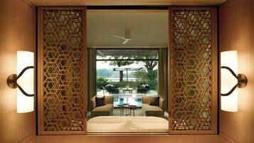 A hotel room with a bed, two armchairs, a coffee table, and a view of an outdoor patio with a pool and umbrellas.