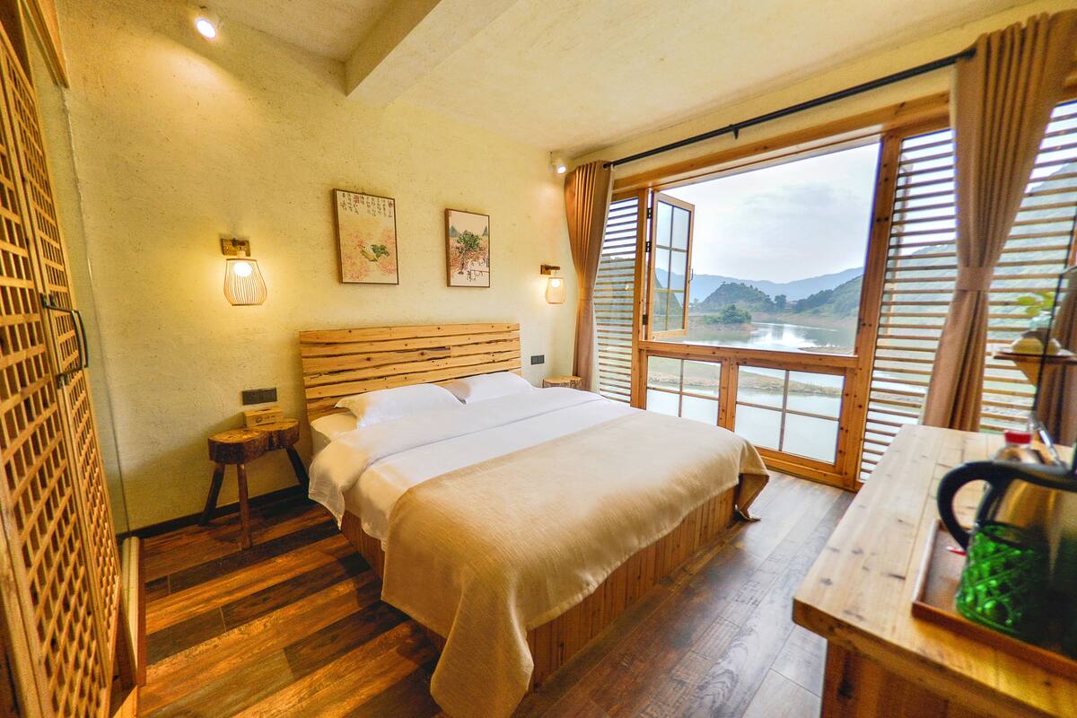 Une chambre avec un lit en bois, une table de chevet, une fenêtre donnant sur un lac et une table avec une bouilloire.