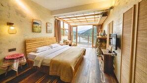 A bedroom with a wooden bed, a small table with a patterned tablecloth, and a view of a mountainous landscape through large windows. - Floral Hotel Qiandaohu Deyishe Inn (Hangzhou)