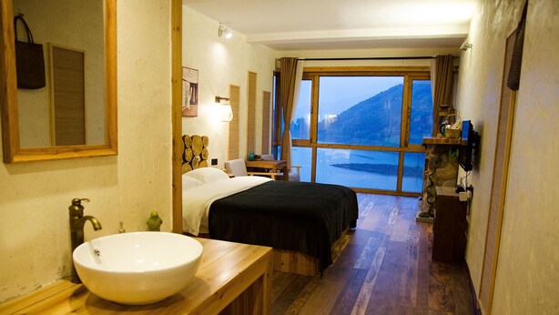 A hotel room with a wooden floor, a large window with a view of a mountain, a bed with a black bedspread, a wooden bedside table, and a white sink. - Floral Hotel Qiandaohu Deyishe Inn (Hangzhou)