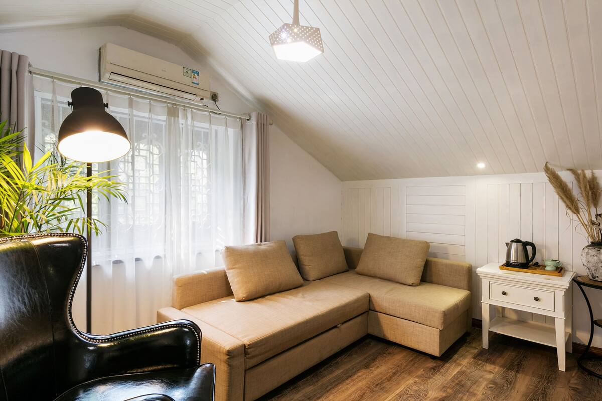 Tunnelmallinen huone, jossa on beige värinen kulmasohva, musta nahkainen nojatuoli, valkoinen yöpöytä, jossa on vedenkeitin, ja ruukussa oleva viherkasvi.