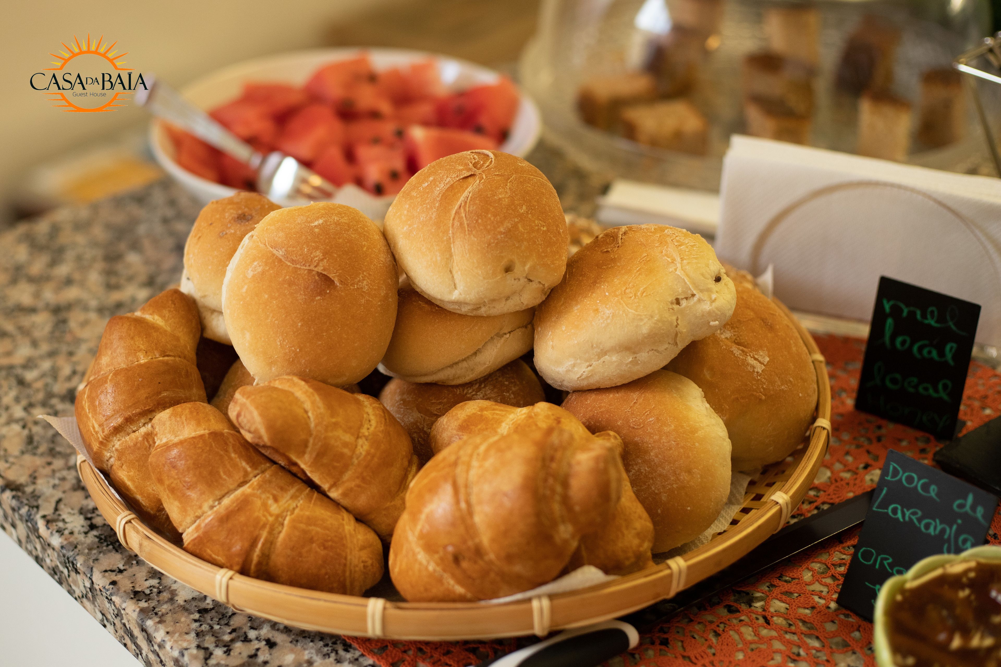 A basket of assorted bread rolls and croissants on a countertop, with a bowl of watermelon slices in the background.