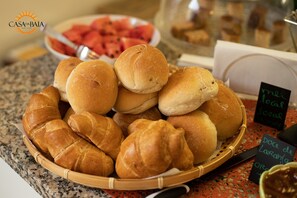 A basket of assorted bread rolls and croissants on a countertop, with a bowl of watermelon slices in the background. - Casa da Baía (Horta)