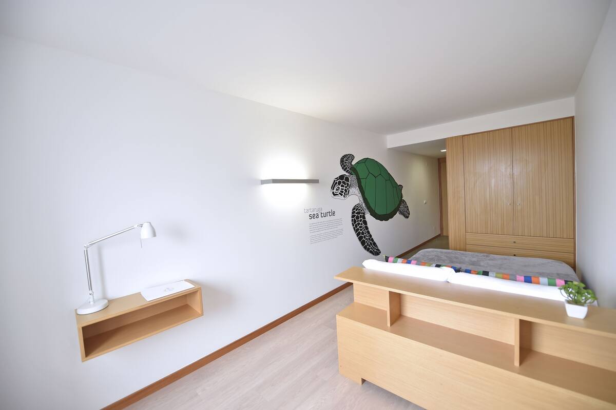 Un dormitorio moderno con una cama de madera, un escritorio con lámpara, un estante en la pared y un mural de tortuga marina.