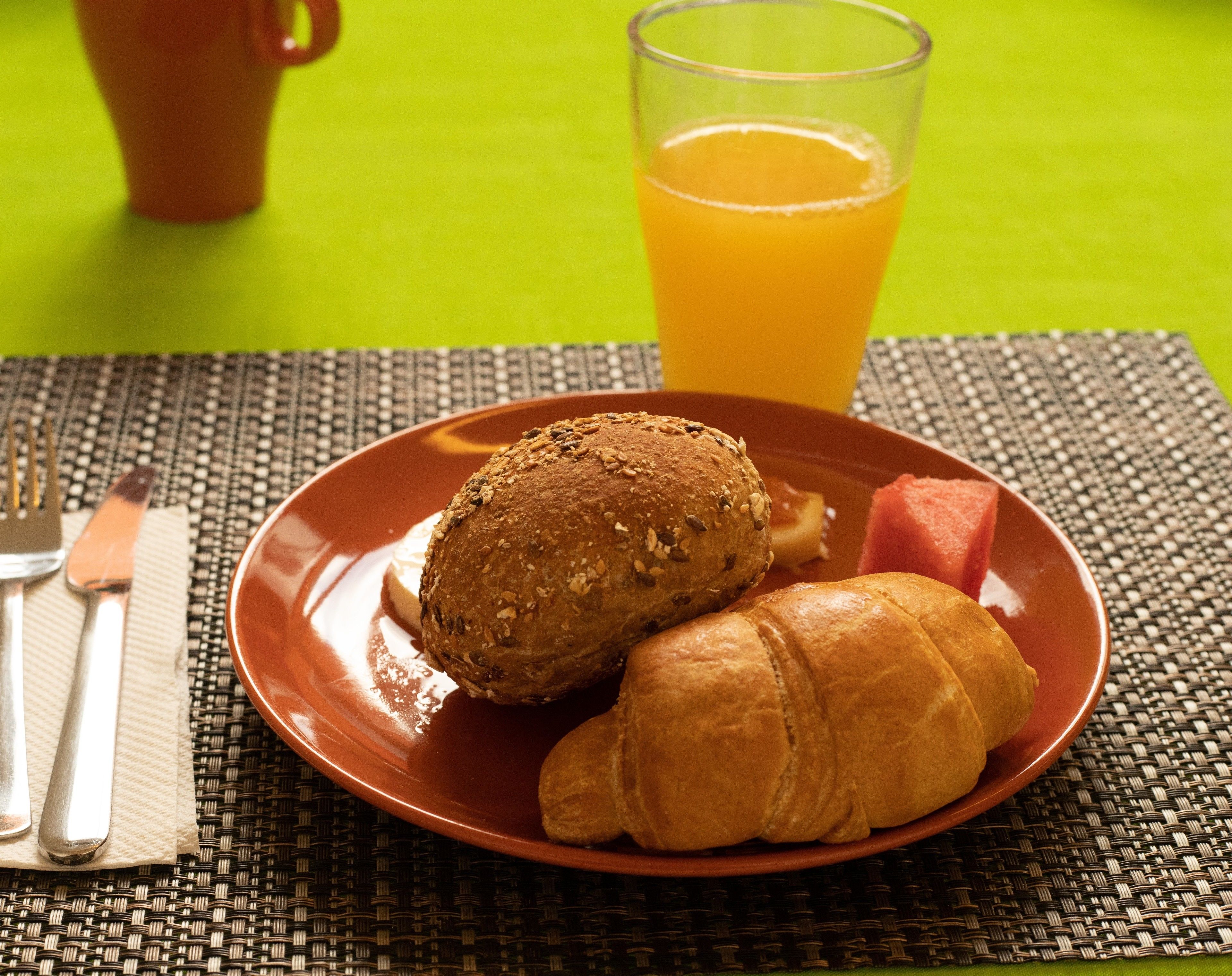 Ein Frühstücksteller mit einem Croissant und einer Scheibe Brot, ein Glas Orangensaft und eine Tasse auf einer grünen Tischdecke.