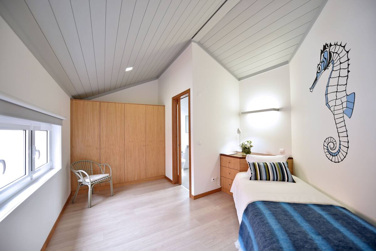 Un dormitorio con un armario de madera, una cama con manta a rayas, un adhesivo mural de caballito de mar y una ventana con persianas.