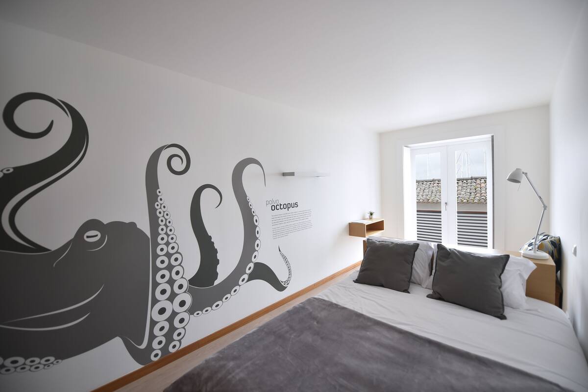 Un dormitorio con un gran mural de pulpo, una cama con ropa de cama gris, una mesita de noche con lámpara y una ventana con contraventanas.
