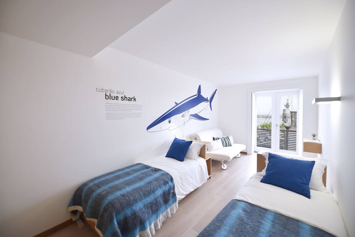 Un dormitorio moderno con dos camas, un vinilo con forma de tiburón azul y un balcón con vistas.